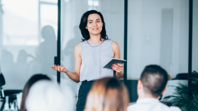 Skolotāja stāv auditorijas priekšā un uzrunā klātesošos