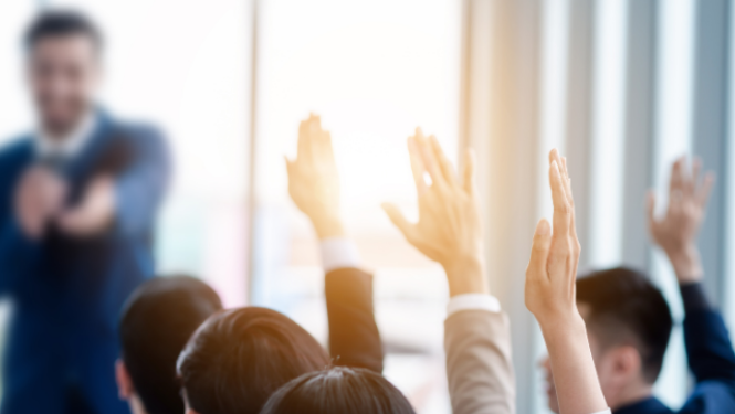 Cilvēki auditorijā paceļ rokas, piedaloties pasākumā vai diskusijā.
