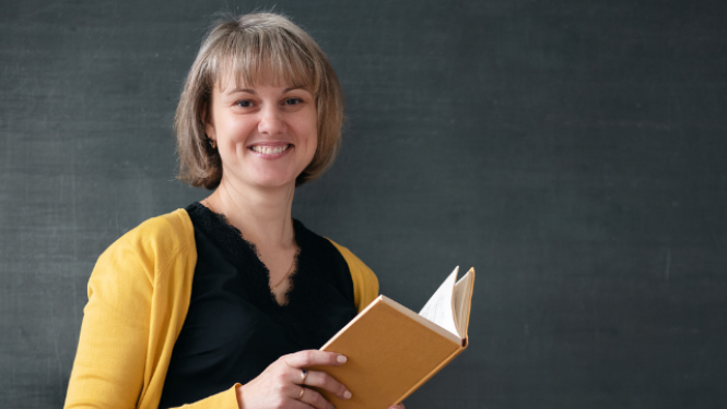 Skolotāja stāv klases priekšā un lasa grāmatu.